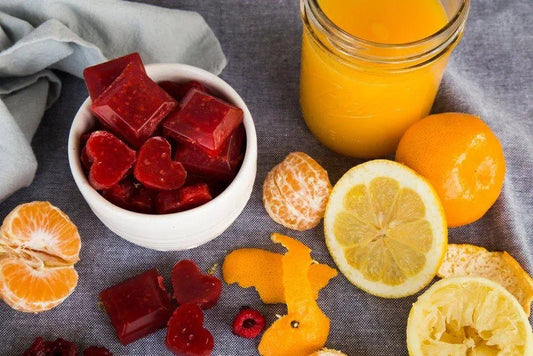 A bowl of Orange and Raspberry Vitamin C gummies surrounded by oranges, lemon slices, orange peel, raspberries and a glass of orange juice 