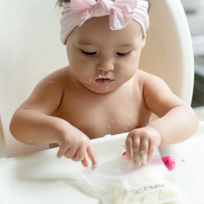 A toddler sits in a highchair, trying to get snacks out of a pouch thats sits on the highchair tray.