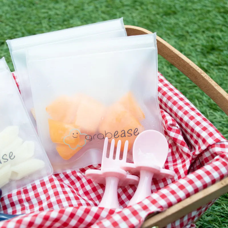 Clear silicone snack pouches filled with rockmelon and pasta sitting in a picnic basket with red and white gingham fabric. A set of blush pink grabease baby fork and spoon sits in the basket in front of the snack pouches. 